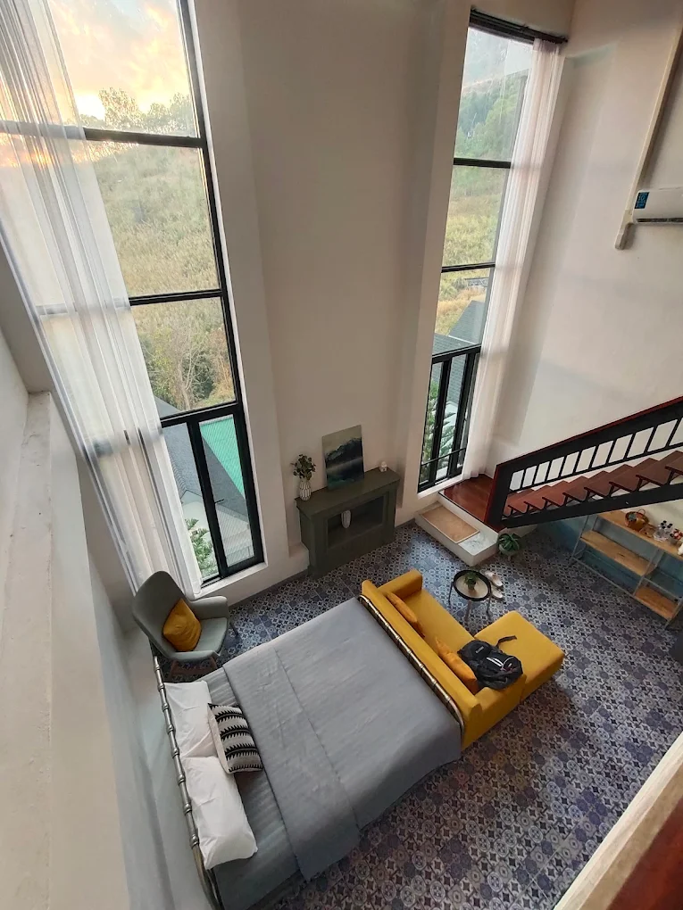 Double-height Spring villa with floor-to-ceiling windows, a grey bed, a yellow bench, and a spiral staircase.